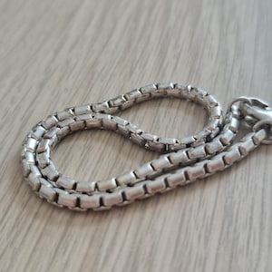 May include: A silver-toned box chain necklace featuring a series of interconnected square links. The chain is arranged in a loose, circular loop and includes a small, circular metal clasp mechanism at one end for securing the piece around the neck.