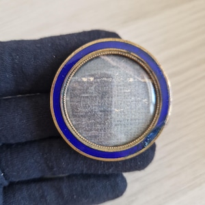 May include: A circular locket featuring a deep blue enamel border set within a gold-toned metal frame. The center contains a glass-covered compartment holding a piece of dark, textured fabric with visible light-colored horizontal lines.