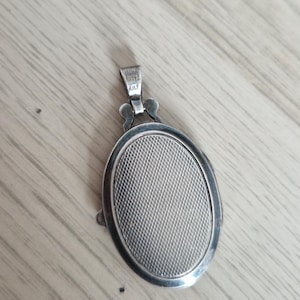 May include: An oval-shaped silver locket pendant featuring a textured, cross-hatched pattern on its front surface. The pendant includes a small, decorative bail at the top with a visible hallmark stamp, and it has a smooth, polished silver border.