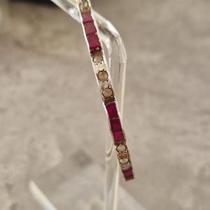 May include: A silver bangle bracelet featuring alternating rectangular red and round clear gemstones. The bracelet is displayed on a clear stand. The gemstones are set in a silver band, highlighting the design.