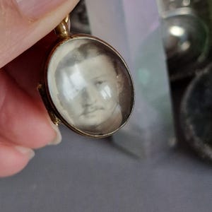 Puede incluir: Un medallón de oro con un frente de vidrio que presenta una foto en blanco y negro de una cara.