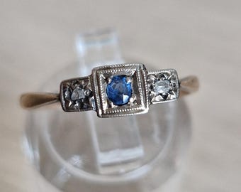 May include: A gold ring featuring a central square setting with a blue oval gemstone, flanked by two smaller square settings each containing a clear gemstone. The metal band has a polished finish and the settings have a milgrain edge detail.