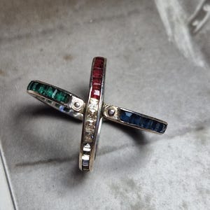 Könnte beinhalten: Ein mehrteiliger Ring bestehend aus zwei ineinandergreifenden Metallbändern. Ein Band ist mit quadratisch geschliffenen grünen und blauen Edelsteinen besetzt. Das andere Band trägt quadratische rote und klare weiße Steine in Kreuzform.