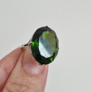 May include: A ring featuring a large, oval-shaped, faceted dark green gemstone set in a silver-toned metal band. The stone displays deep forest green hues with reflective surfaces and is secured by four small prongs at the top, bottom, and sides.