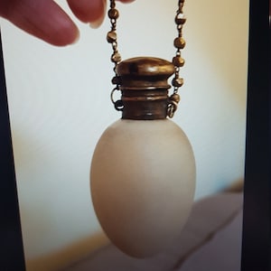 Könnte beinhalten: Eine mattierte Glasflasche mit einem goldenen Metallverschluss und einer Kette. Die Flasche ist oval geformt und hängt an einer Kette.