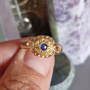 Könnte beinhalten: Ein goldfarbener Ring mit einem verzierten Design. Der Ring hat einen zentralen blauen Edelstein, umgeben von detaillierten Filigranarbeiten und dekorativen Elementen. Das Band hat ein strukturiertes Aussehen.