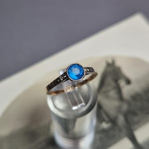 Puede incluir: Un anillo vintage con una piedra preciosa azul brillante engastada en una banda plateada. La banda presenta pequeñas piedras de acento transparentes y un interior dorado. El anillo se exhibe sobre un soporte transparente sobre un fondo borroso.