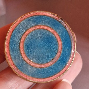 May include: A circular brooch featuring a gold-toned metal frame with concentric rings of textured light blue enamel and salmon pink enamel. The surface displays a fine, wavy guilloché pattern beneath the translucent blue enamel layers.