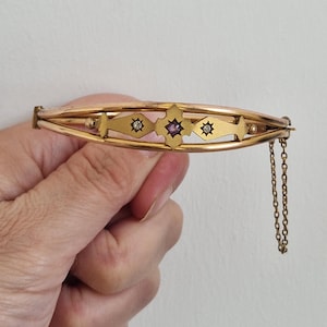 Puede incluir: Un brazalete rígido articulado de tono dorado con una barra decorativa central con un engaste en forma de estrella. El centro contiene una pequeña piedra preciosa morada flanqueada por dos piedras claras, cada una engastada en un motivo de estrella.