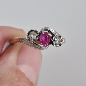 May include: A ring featuring a central oval-cut pinkish-red gemstone flanked by two round-cut clear stones. The setting is crafted from a combination of yellow and white metal, with intricate engraved detailing along the band and stone mounts.