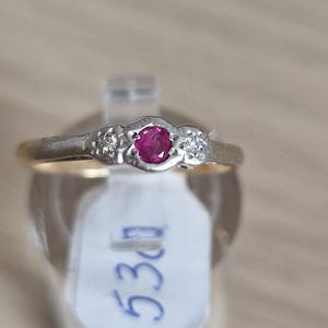 May include: A gold ring featuring a central round red ruby flanked by two small round clear diamonds set in a white metal mounting. The ring is displayed on a clear stand with a white tag attached that has the handwritten blue text 530.