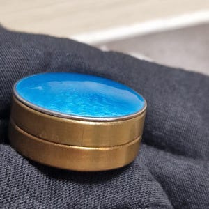 Op de afbeelding: Een kleine, ronde, antieke sieradendoos met een helderblauwe emaille deksel en een goudkleurige metalen basis. De deksel heeft een zilverkleurige metalen rand. De doos wordt waarschijnlijk gebruikt voor het opbergen van kleine items zoals sieraden of pillen.