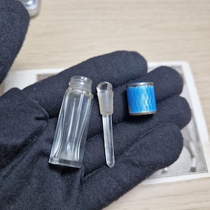 May include: A small, clear glass perfume bottle with a matching glass stopper and a blue and silver container. The bottle has a rectangular base and a narrow, ridged body. The blue container has a silver rim.