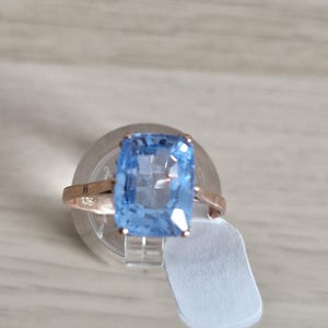May include: A ring featuring a large, rectangular, light blue faceted gemstone set in a rose gold band. The band has a visible hallmark stamp on the side and is attached to a small white rectangular price tag, resting on a clear circular stand.