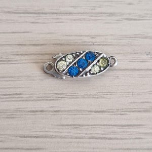 Può includere: Una chiusura per gioielli di forma ovale con una montatura in metallo color argento e un motivo decorativo. Il centro è impreziosito da tre file diagonali di piccole pietre rotonde sfaccettate, tra cui due file di pietre chiare e una blu.