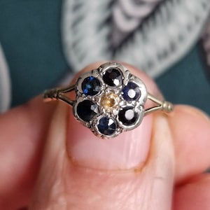 May include: A silver ring with a flower-shaped setting featuring six blue sapphires and a single yellow stone in the center.