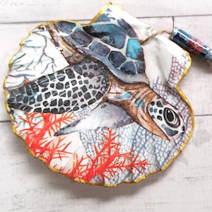 May include: A decorative seashell with a painted sea turtle design. The artwork features shades of blue, brown, and white, with a gold-painted edge and a jute string with a wooden bead attached.