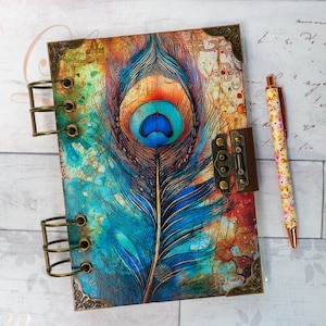 Pode incluir: Um diário com um design vibrante de pena de pavão. A capa apresenta uma pena grande e detalhada em tons de azul, laranja e verde-azulado. Possui um fecho de metal e protetores de canto. Uma caneta floral repousa ao lado do diário.