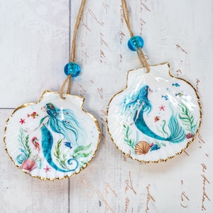 May include: Two seashell ornaments, each featuring a mermaid illustration with blue hair and tail, surrounded by aquatic plants and fish. The shells have a gold-painted edge and are suspended by twine and blue beads.