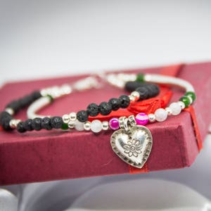 May include: A silver heart charm with a floral design hangs from a black lava rock and white bead bracelet. The bracelet is displayed in a red gift box.