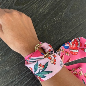 Könnte beinhalten: Ein rosa-grüner Schal mit floralem Muster und einem goldenen Ringakzent. Der Schal ist als Armband um ein Handgelenk gebunden.