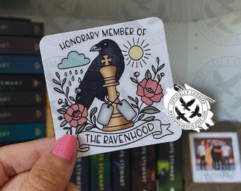 May include: A square sticker featuring a black raven perched on a gold chess king piece, flanked by pink flowers, a blue cloud with raindrops, and a yellow sun. The text reads HONORARY MEMBER OF THE RAVENHOOD on a white background.