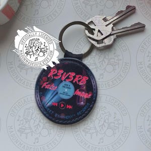 May include: A circular black keychain featuring a graphic design of a vinyl record with the text R3V3RB and False Image printed in pink. The keychain is attached to a metal ring holding two silver keys. The design includes a microphone icon.