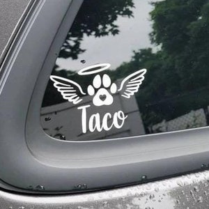 May include: A white vinyl decal with a paw print inside a halo and wings, with the name "Taco" underneath. The decal is on the back window of a gray car.