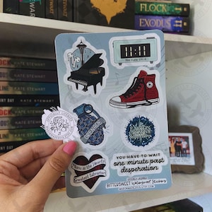 Peut inclure: Une planche d'autocollants illustrée d'un piano à queue noir, d'une horloge numérique affichant 11:11, d'une basket montante rouge, d'une grenade bleue, d'une étoile et d'un cœur. Le texte inclut "Make a wish, Stella", "Easton Crowne Supernova", "Music is the heart's greatest librarian" et "You have to wait one minute past desperation".