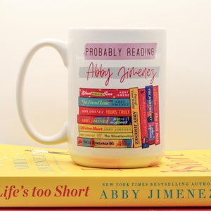 May include: White ceramic mug with the text "Probably Reading" and "Abby Jimenez" in pink and gray. The mug features a stack of colorful book spines. The mug is sitting on top of two books.