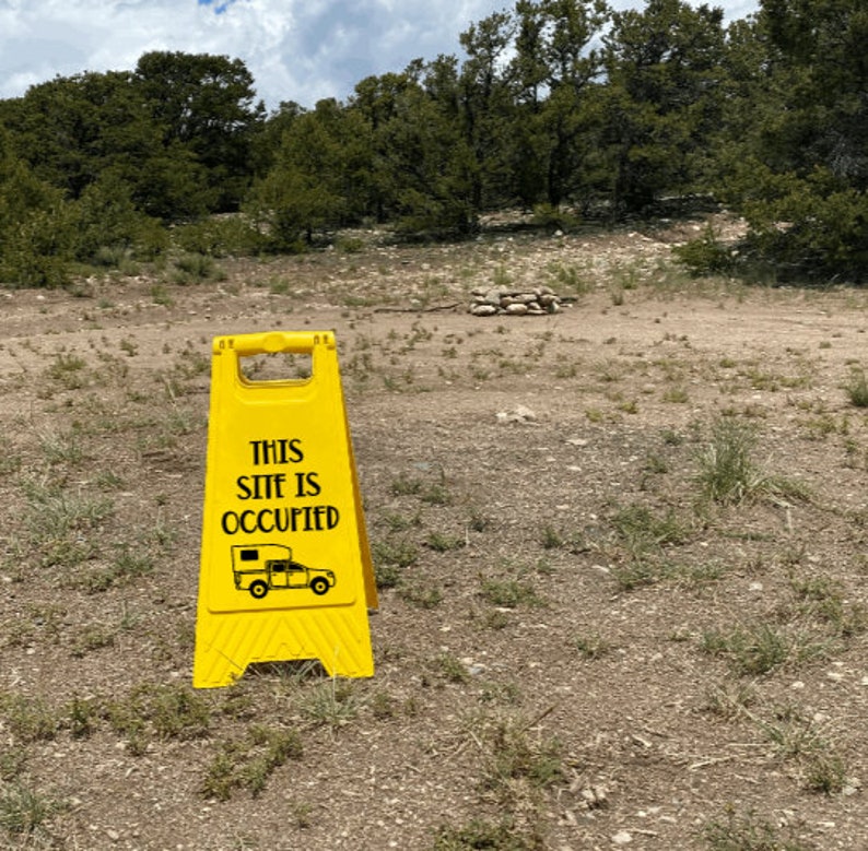May include: A yellow caution sign with black text that reads "This Site Is Occupied" and a black silhouette of a camper van. The sign is on a dirt road with trees in the background.
