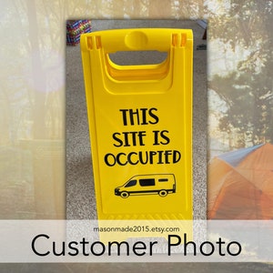 May include: Yellow camping sign with black text that reads "This Site Is Occupied" and a black silhouette of a camper van. The sign is on a gray surface with a yellow tent in the background.