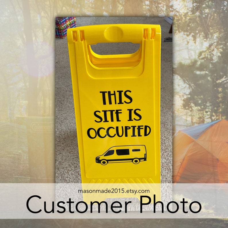 May include: Yellow camping sign with black text that reads "This Site Is Occupied" and a black silhouette of a camper van. The sign is on a gray surface with a yellow tent in the background.