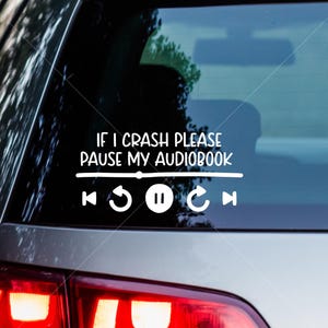 May include: White vinyl car decal with the text "IF I CRASH PLEASE PAUSE MY AUDIOBOOK" above a music player interface. The interface includes rewind, play/pause, and fast forward icons. The decal is applied to a car window.