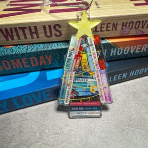 May include: A clear acrylic Christmas tree ornament featuring a yellow star and a stack of book covers. The book titles include "Ugly Love" and "Maybe Now" by Colleen Hoover. The ornament is displayed in front of a stack of books.
