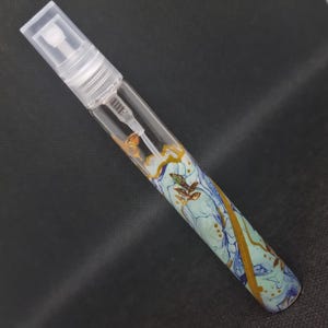 May include: A clear glass perfume atomizer with a decorative painted design. The design features swirls of blue, gold, and green. The atomizer has a clear plastic spray nozzle and cap.