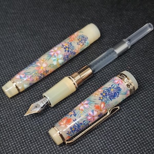 May include: A fountain pen with a cream-colored body featuring a colorful floral pattern of pink, orange, and blue flowers. The pen includes a gold-toned metal clip, a silver-toned nib with intricate engravings, and a clear plastic ink converter.