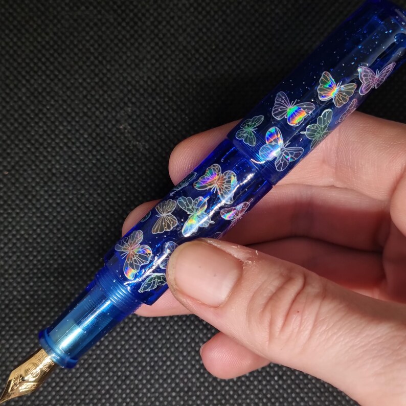 Puede incluir: Una pluma estilogr&aacute;fica azul con plum&iacute;n dorado, adornada con dise&ntilde;os de mariposas iridiscentes. El cuerpo de la pluma es azul transl&uacute;cido, y las mariposas brillan con colores arco&iacute;ris. La pluma se sostiene en una mano sobre un fondo oscuro.