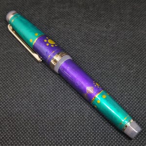 K&ouml;nnte beinhalten: Dekorativer F&uuml;llfederhalter mit einem t&uuml;rkis-violetten Schaft. Der Stift hat goldfarbene Akzente und einen Clip. Der Schaft ist mit gelben Punkten und geometrischen Formen verziert. Der Stift liegt auf einer dunkelgrauen Oberfl&auml;che.