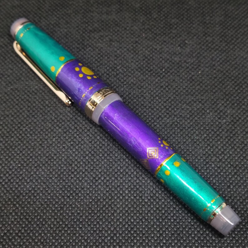 K&ouml;nnte beinhalten: Dekorativer F&uuml;llfederhalter mit einem t&uuml;rkis-violetten Schaft. Der Stift hat goldfarbene Akzente und einen Clip. Der Schaft ist mit gelben Punkten und geometrischen Formen verziert. Der Stift liegt auf einer dunkelgrauen Oberfl&auml;che.