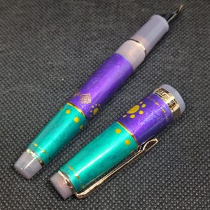 K&ouml;nnte beinhalten: Ein zerlegter F&uuml;llfederhalter mit einem t&uuml;rkis-violetten Schaft. Der Stift hat goldene Akzente und einen goldenen Clip. Der Schaft ist mit gelben Punkten und einem geometrischen Design verziert. Die Feder des Stifts ist sichtbar.