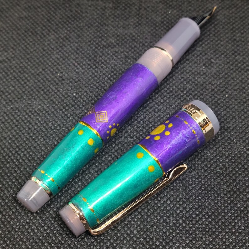 K&ouml;nnte beinhalten: Ein zerlegter F&uuml;llfederhalter mit einem t&uuml;rkis-violetten Schaft. Der Stift hat goldene Akzente und einen goldenen Clip. Der Schaft ist mit gelben Punkten und einem geometrischen Design verziert. Die Feder des Stifts ist sichtbar.