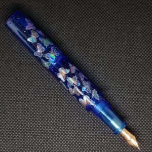 Puede incluir: Pluma estilogr&aacute;fica azul con plum&iacute;n dorado. El cuerpo de la pluma est&aacute; decorado con dise&ntilde;os de mariposas iridiscentes. La pluma est&aacute; hecha de un material transparente y brillante. El capuch&oacute;n no es visible en la imagen.