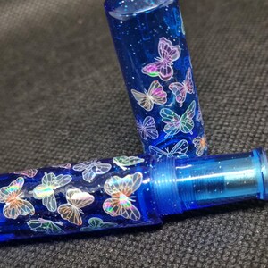 Puede incluir: Un bol&iacute;grafo azul con una tapa transparente, adornado con dise&ntilde;os de mariposas iridiscentes. El cuerpo y la tapa del bol&iacute;grafo est&aacute;n llenos de purpurina, creando un efecto brillante. Las mariposas tienen un brillo hologr&aacute;fico.