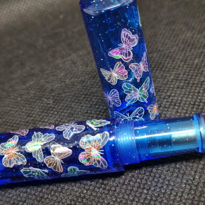 Puede incluir: Un bol&iacute;grafo azul con una tapa transparente, adornado con dise&ntilde;os de mariposas iridiscentes. El cuerpo y la tapa del bol&iacute;grafo est&aacute;n llenos de purpurina, creando un efecto brillante. Las mariposas tienen un brillo hologr&aacute;fico.