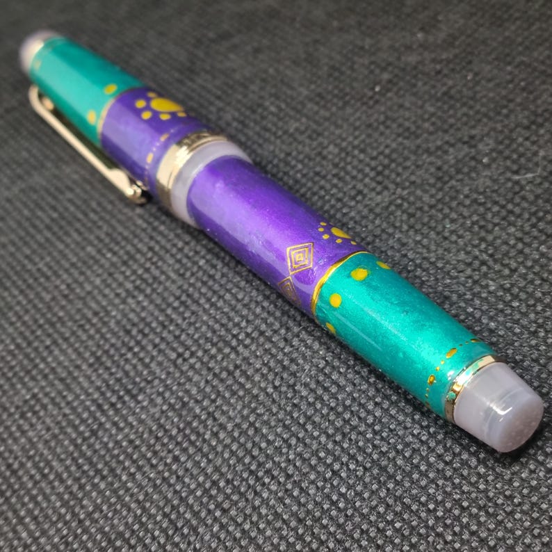 K&ouml;nnte beinhalten: Ein farbenfroher Stift mit t&uuml;rkis, lila und goldenem Design. Der Stift hat einen goldenen Clip und Akzente sowie eine transparente Spitze. Der Stiftschaft weist ein Muster aus gelben Punkten und geometrischen Formen auf.