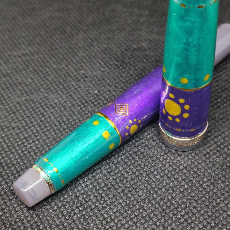 K&ouml;nnte beinhalten: Ein dekorativer Stift mit einem t&uuml;rkis-violetten Schaft. Der Stift hat goldfarbene Akzente und ein geometrisches Design. Die Kappe des Stifts ist abgenommen und gibt die Feder frei. Der Stift liegt auf einer strukturierten, dunkelgrauen Oberfl&auml;che.