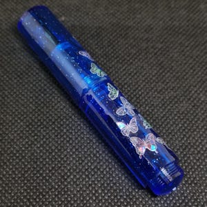 Puede incluir: Un bol&iacute;grafo azul con un dise&ntilde;o brillante y moteado y calcoman&iacute;as de mariposas. El bol&iacute;grafo tiene un cuerpo transparente y una tapa de rosca. Las mariposas son blancas con acentos iridiscentes.