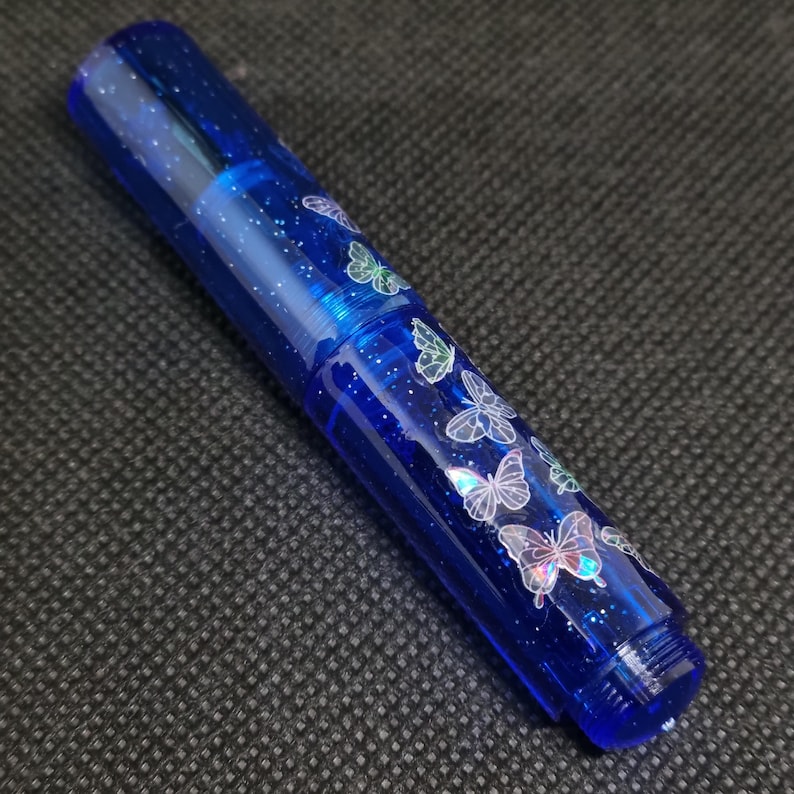 Puede incluir: Un bol&iacute;grafo azul con un dise&ntilde;o brillante y moteado y calcoman&iacute;as de mariposas. El bol&iacute;grafo tiene un cuerpo transparente y una tapa de rosca. Las mariposas son blancas con acentos iridiscentes.