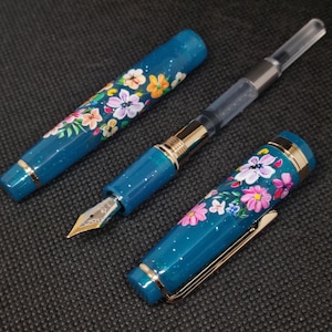 May include: A teal fountain pen featuring a floral pattern. The pen's body and cap are decorated with hand-painted flowers in pink, orange, and white. Gold-coloured trim and a gold nib are also visible. The pen is shown in pieces.